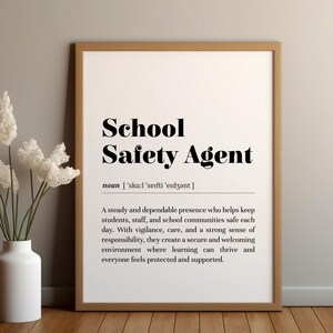 Puede incluir: Una impresión enmarcada con las palabras "School Safety Agent" en negrita negra. La definición del término está debajo del título. Un jarrón blanco con flores blancas está a la izquierda de la impresión enmarcada.