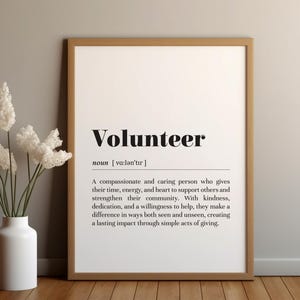 以下が含まれることがあります： 太字の黒い文字で「Volunteer」という言葉とその定義が書かれた額入りのプリント。プリントの左側には、白い花が入った白い花瓶が置かれています。