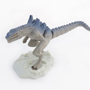 Könnte beinhalten: Ein graues und blaues Plastik-Dinosaurierspielzeug mit einem langen Schwanz und scharfen Zähnen. Der Dinosaurier steht auf einem weißen Sockel.