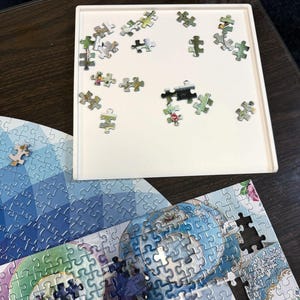 May include: A white puzzle tray with scattered puzzle pieces. The pieces are various colors and patterns, some with floral designs. Partially assembled puzzles with blue and green imagery are visible below the tray.