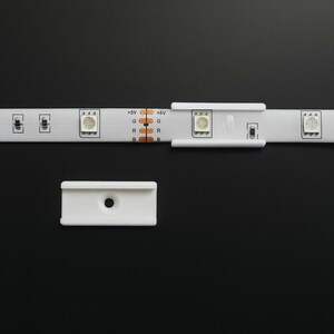 May include: A white LED strip with multiple LED lights and a white plastic connector. The strip has markings for +5V, G, R, and B. A separate white plastic clip is also visible.