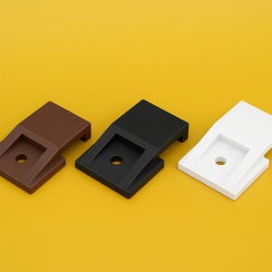 Puede incluir: Tres soportes de plástico en marrón, negro y blanco se disponen sobre un fondo amarillo. Cada soporte tiene una base rectangular con un orificio central y una sección superior inclinada elevada.