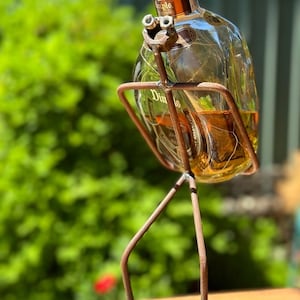 Könnte beinhalten: Eine durchsichtige Glasflasche mit Dimple Scotch Whisky steht in einem Vogel-förmigen Metallständer. Der Ständer hat zwei Beine und eine runde Basis. Die Flasche ist teilweise mit bernsteinfarbener Flüssigkeit gefüllt.