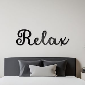 May include: A black metal wall decor with the word "Relax" in a cursive font, above a gray upholstered bed with white bedding and two pillows.