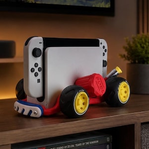 May include: A white and black Nintendo Switch console is mounted on a red and black car-shaped stand with yellow wheels. The stand has the word "FUEL" printed on it. The console is on a wooden table with video game cases below.