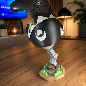 May include: A black and white Chain Chomp controller stand holding a black game controller. The stand features a spherical character head with white eyes, a gray articulated chain base, and a tan hexagonal platform with small green bushes.