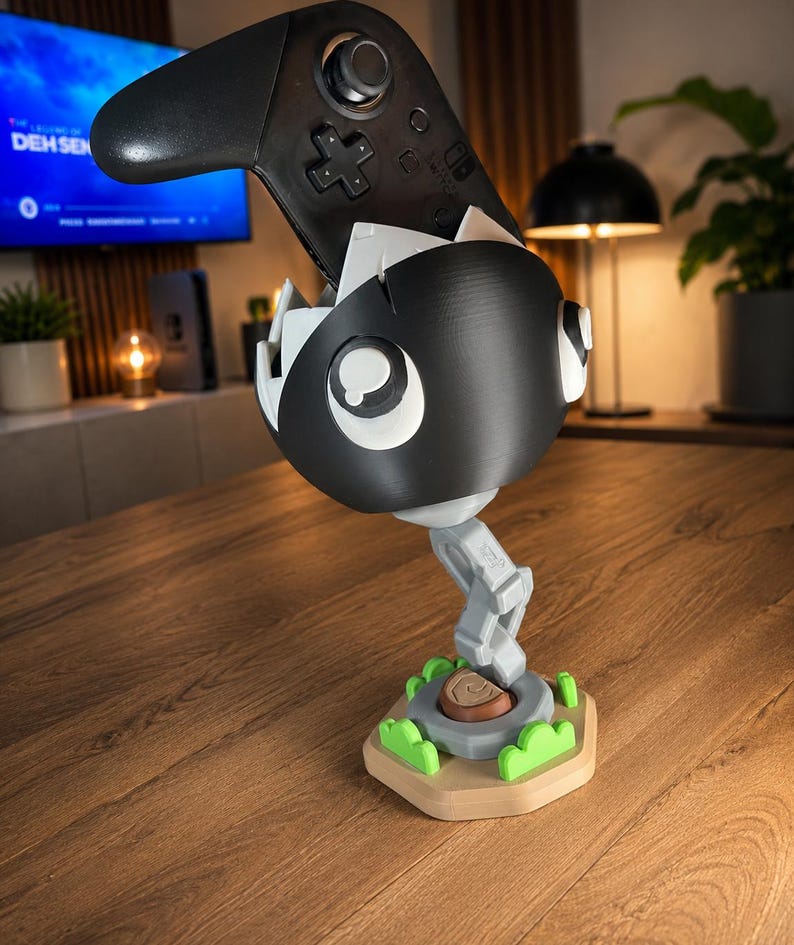 May include: A black and white Chain Chomp controller stand holding a black game controller. The stand features a spherical character head with white eyes, a gray articulated chain base, and a tan hexagonal platform with small green bushes.