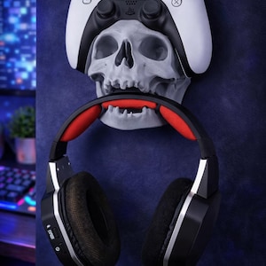 Puede incluir: Una configuración de juego con un mando de PlayStation 5 blanco y negro apoyado en una calavera gris, con auriculares negros y rojos colgando debajo. El fondo es azul oscuro.