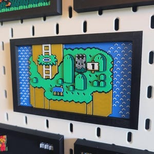Op de afbeelding: Ingelijste pixelkunst die een klassiek videogamelevel uitbeeldt. Het kunstwerk toont een groen landschap met een kasteel, water en het nummer 1. De lijst is zwart en de kunst is gemonteerd op een wit pegboard.