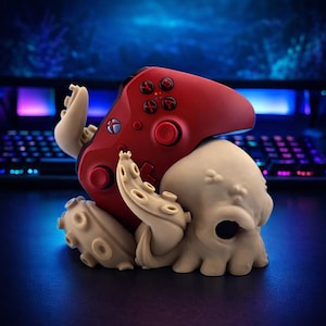 May include: A red video game controller rests on a beige octopus-shaped stand. The controller has black buttons and a directional pad. The stand has detailed tentacles and a skull-like head, creating a unique gaming accessory.