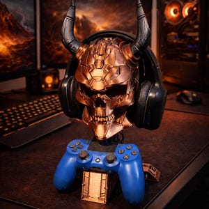 Puede incluir: Un soporte para auriculares con forma de calavera de color cobre con cuernos y auriculares negros. Un mando de juego azul descansa sobre un soporte debajo de la calavera. El soporte está sobre una superficie marrón oscuro, con monitores de ordenador y un teclado en el fondo.