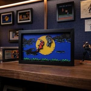 Puede incluir: Una pieza de arte en caja de sombra rectangular que presenta una escena 3D en capas de un ninja rojo y un ninja azul saltando frente a una gran luna llena amarilla contra un cielo azul oscuro con nubes negras y follaje verde, enmarcada en negro.