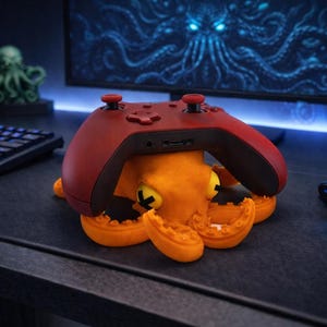 May include: An orange octopus-shaped controller holder with a red controller resting on top. The octopus has yellow eyes with black cross marks. A green octopus figurine and a computer keyboard are in the background.