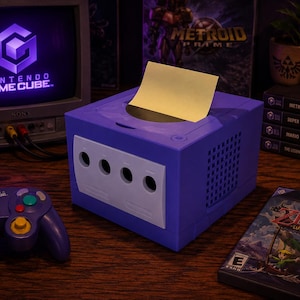 May include: A purple and white square tissue box cover designed to resemble a vintage video game console. The box features a front panel with four circular controller ports and a top opening where a yellow rectangular paper sheet is inserted.