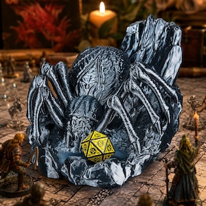 May include: A detailed, dark gray spider-shaped dice holder with a yellow and black twenty-sided die. The spider is surrounded by miniature figurines, creating a fantasy gaming scene. A lit candle and a "Dungeons & Dragons" book are in the background.