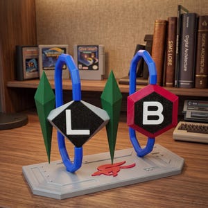 Op de afbeelding: Een decoratief displaystuk met twee blauwe zeshoekige ringen op een grijze rechthoekige basis. Twee groene ruitvormige prisma's flankeren de ringen. De ene ring bevat een zwarte ruit met de letter L, de andere een rode zeshoek met de letter B.
