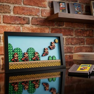 Puede incluir: Un marco rectangular negro tipo caja que contiene un diorama 3D multicapa de una escena de videojuego de Super Mario. La escena presenta personajes pixelados, colinas verdes, una moneda amarilla y un patrón de suelo texturizado.