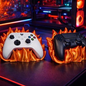 May include: A white Xbox controller and a black PlayStation controller rest on orange flame-shaped stands. The controllers are on a dark surface, with a computer and keyboard in the background. The scene is lit with red and blue lights.
