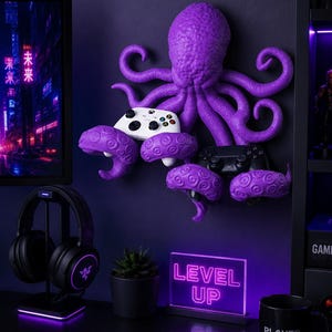 May include: A purple octopus-shaped wall mount holding two game controllers. A white controller is in the center, and a black controller is on the right. A neon sign that says "LEVEL UP" is on the desk, along with a black mug that says "PLAYER ONE".