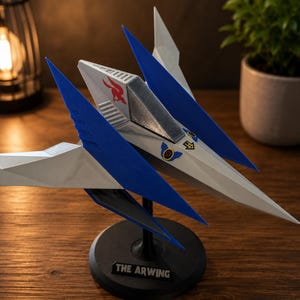 Könnte beinhalten: Ein weiß-blaues Modell eines Sternenjägers, montiert auf einem schwarzen, runden Präsentationsständer. Das Raumschiff hat ein rotes Vogel-Emblem, ein silbernes Cockpit und kleine Aufkleber. Eine schwarze Plakette am Sockel trägt den Text THE ARWING.