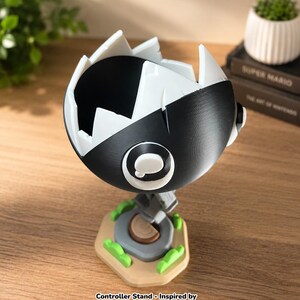 May include: A black and white spherical controller stand featuring a jagged, tooth-like opening at the top. The stand is mounted on a tan hexagonal base with small green accents and a central gray circular element. Text reads: Controller Stand - Inspired by Mario's Chain Chomp.
