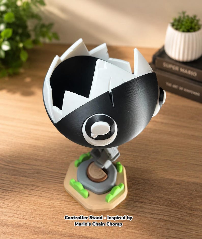 May include: A black and white spherical controller stand featuring a jagged, tooth-like opening at the top. The stand is mounted on a tan hexagonal base with small green accents and a central gray circular element. Text reads: Controller Stand - Inspired by Mario's Chain Chomp.