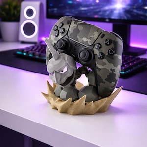 May include: A camouflage-patterned video game controller rests on a gray and tan decorative stand. The stand features a stylized, gray, cartoon-like character. The controller has black buttons and directional controls. The background includes a computer monitor and keyboard.