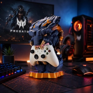 May include: A blue and gold Predator-themed gaming controller holder. The holder features a detailed sculpted head design, holding a white Xbox controller. The base is gold, and the background includes a computer monitor and gaming PC.