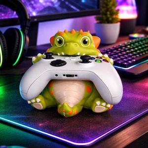 以下が含まれることがあります： 白いゲームコントローラーを抱える、緑とオレンジ色の恐竜型コントローラーホルダー。 ホルダーには大きな目があり、黒い表面にあります。 コンピューターモニター、キーボード、マウスが背景にあります。