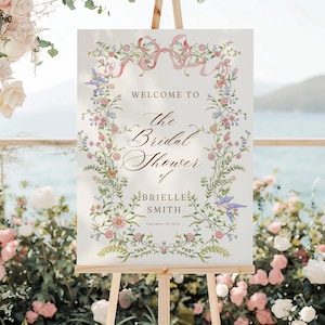 Könnte beinhalten: Ein Willkommensschild für eine Brautparty, das auf einer Holzstaffelei präsentiert wird. Das Schild zeigt florale und Bandakzente mit dem Text "Welcome to the Bridal Shower of Brielle Smith". Der Hintergrund zeigt Blumen und eine malerische Aussicht.