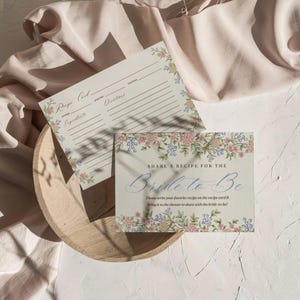 May include: Two recipe cards with floral borders rest on a wooden tray. One card is a recipe template, the other reads "Share a Recipe for the Bride-to-Be." The cards feature pink, blue, and green floral designs.