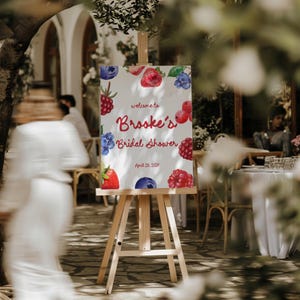 May include: A wooden easel displays a sign for "Brooke's Bridal Shower" with watercolor illustrations of berries. The sign's text is in red and the date is April 29, 2023. The background shows an outdoor event.