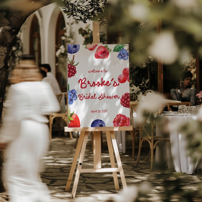 May include: A wooden easel displays a sign for "Brooke's Bridal Shower" with watercolor illustrations of berries. The sign's text is in red and the date is April 29, 2023. The background shows an outdoor event.