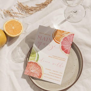 May include: A bridal shower invitation featuring watercolor citrus fruit slices in orange, pink, and lime green. The invitation reads "She found her Main Squeeze" and includes event details. The invitation is on a plate with a glass and orange slices.