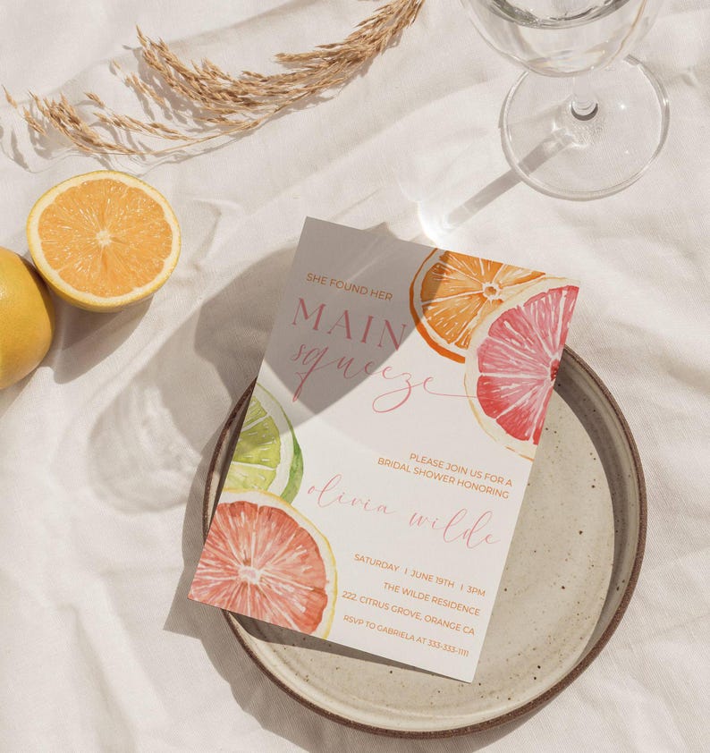 May include: A bridal shower invitation featuring watercolor citrus fruit slices in orange, pink, and lime green. The invitation reads "She found her Main Squeeze" and includes event details. The invitation is on a plate with a glass and orange slices.