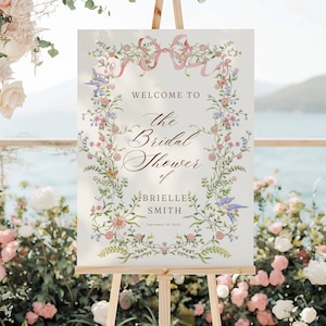 May include: A welcome sign for a bridal shower, displayed on a wooden easel. The sign features floral and ribbon accents, with text that reads "Welcome to the Bridal Shower of Brielle Smith, September 18, 2024."