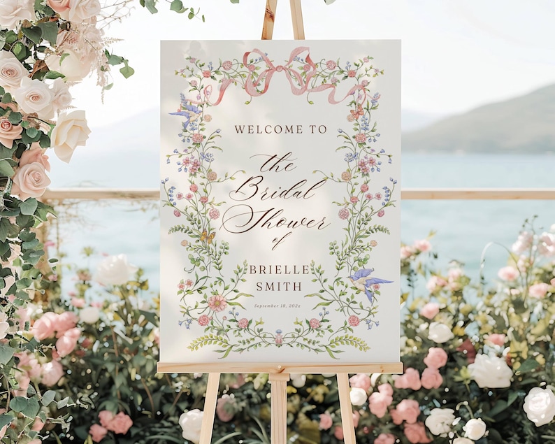 May include: A welcome sign for a bridal shower, displayed on a wooden easel. The sign features floral and ribbon accents, with text that reads "Welcome to the Bridal Shower of Brielle Smith, September 18, 2024."