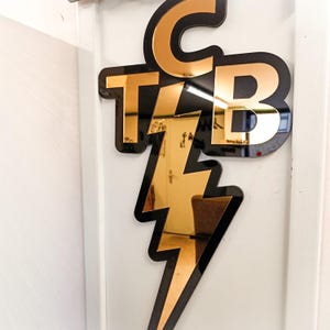 Puede incluir: Un letrero decorativo de pared con las letras "CTB" en acrílico dorado espejado, contorneado en negro, y un rayo dorado. El letrero está montado en una puerta blanca.