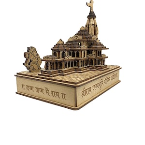 May include: A wooden model of the Shri Ram Janmabhoomi Mandir in Ayodhya, India. The model is intricately detailed and features a golden spire. The base of the model is inscribed with the text "Jai Krishna Krishna Me Ram Ra"