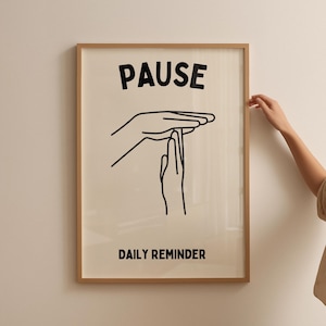 May include: A framed art print featuring the word "PAUSE" in black above a black line drawing of hands in a time-out gesture. The text "DAILY REMINDER" is below the hands. The frame is light brown.