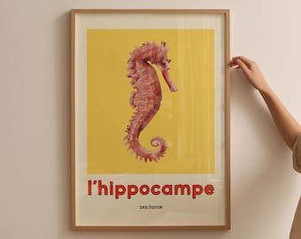 May include: Framed art print featuring a detailed illustration of a seahorse in shades of pink and red against a yellow background. The text "l'hippocampe sea horse" is displayed in red at the bottom of the print. The frame is a light brown color.