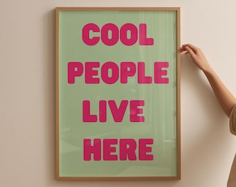 May include: A framed art print with the words "COOL PEOPLE LIVE HERE" in bright pink letters on a light green background. The frame is a light brown color. The print is hanging on a white wall.
