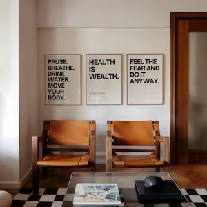 May include: Two wooden armchairs with tan leather seats are positioned below three framed posters with motivational text. The posters read: "PAUSE. BREATHE. DRINK WATER. MOVE YOUR BODY.", "HEALTH IS WEALTH.", and "FEEL THE FEAR AND DO IT ANYWAY."