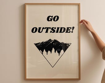 May include: Framed art print with the words "GO OUTSIDE!" in black, above a black and white mountain and tree graphic. The print is in a light wood frame and is displayed on a neutral wall.
