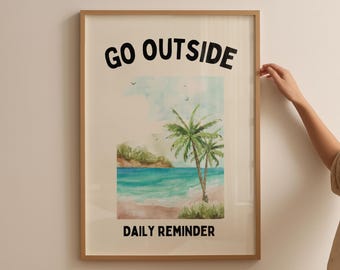 May include: Framed art print with the words "GO OUTSIDE" and "DAILY REMINDER" in black. The artwork features a watercolor beach scene with palm trees, blue water, and a sandy shore. The frame is a light brown color.