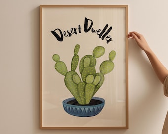 May include: A framed art print featuring a watercolor illustration of a cactus in a blue pot. The artwork is labeled "Desert Dweller" in black script. The cactus is green with yellow accents, set against a cream background, and framed in a light brown wood.