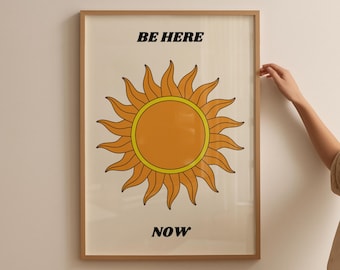 May include: Framed art print featuring a retro-style sun illustration in orange and yellow, with the words "BE HERE NOW" in black text. The artwork is displayed in a light brown wooden frame.