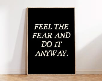 May include: A framed black poster with the text "FEEL THE FEAR AND DO IT ANYWAY" in a bold, white, serif font. The poster is set against a white wall and rests on a wooden floor.