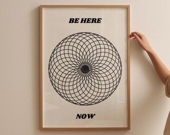 May include: Framed art print with a geometric black and white design. The artwork features a circular pattern and the words "BE HERE NOW" in bold, black font. The frame is a light brown color.