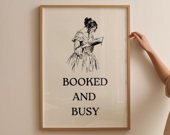 May include: A framed print with a light beige background features a black and white illustration of a woman reading a book. The text "BOOKED AND BUSY" is printed in bold, black letters below the illustration. The frame is a light brown color.
